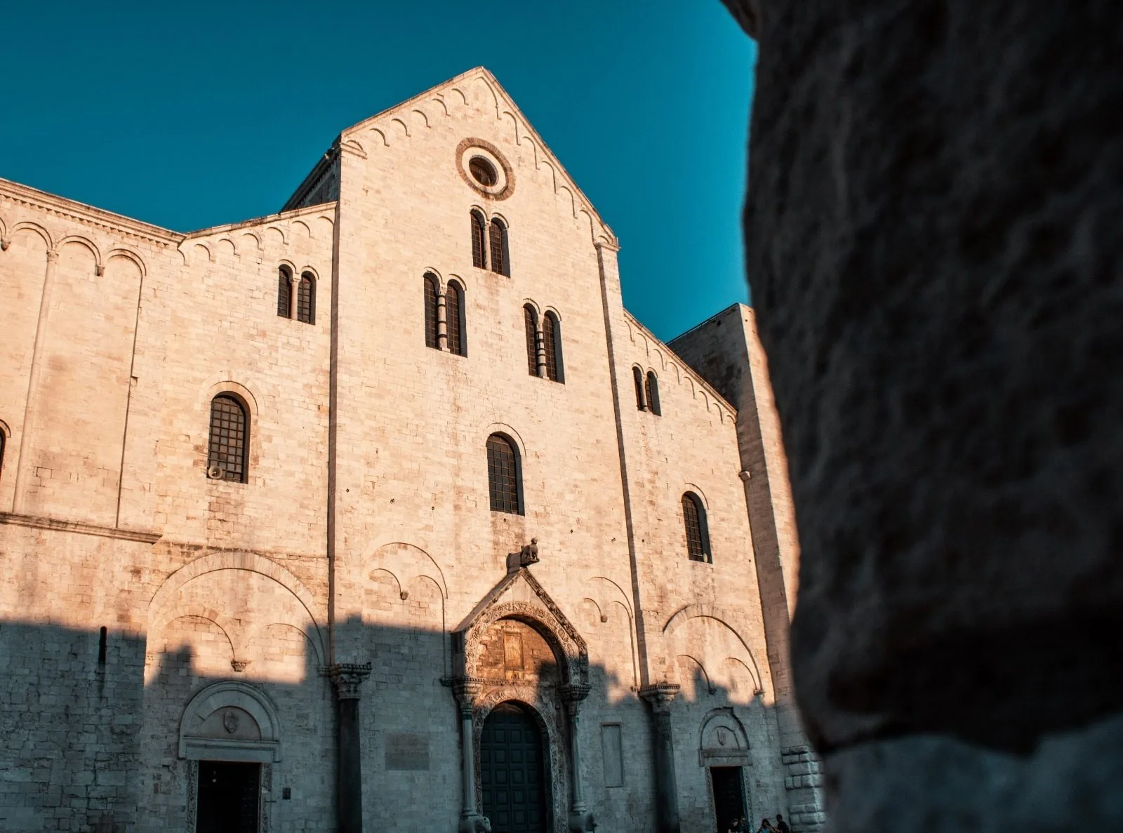 La facciata in pietra chiara della Basilica di San Nicola a Bari illuminata dal sole del tardo pomeriggio, con il portale centrale e le bifore, sotto un cielo blu intenso