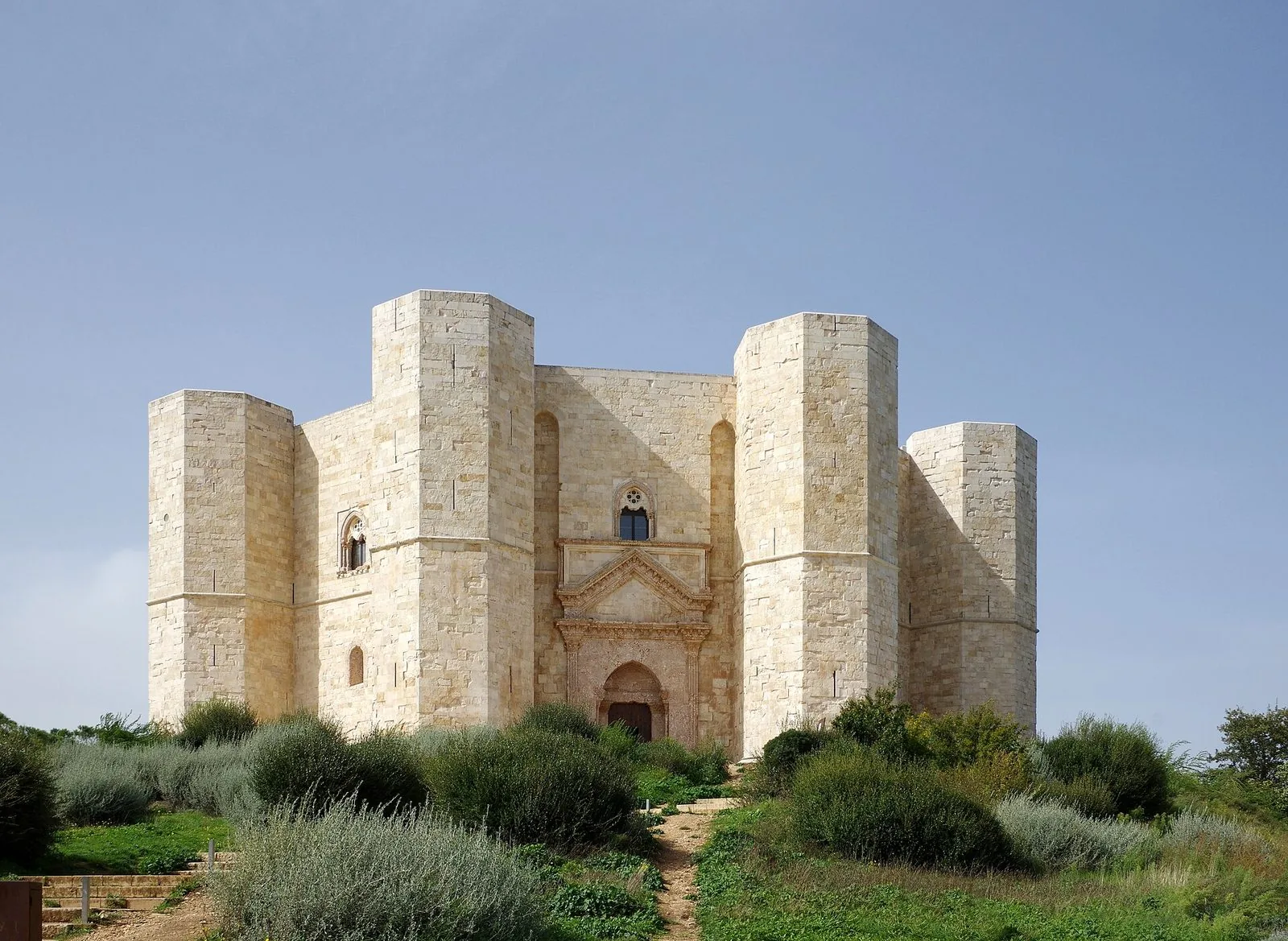Castel del Monte visto dal sentiero di accesso: la facciata in pietra chiara con il portale d'ingresso e le torri ottagonali, tra cespugli di macchia sotto un cielo azzurro