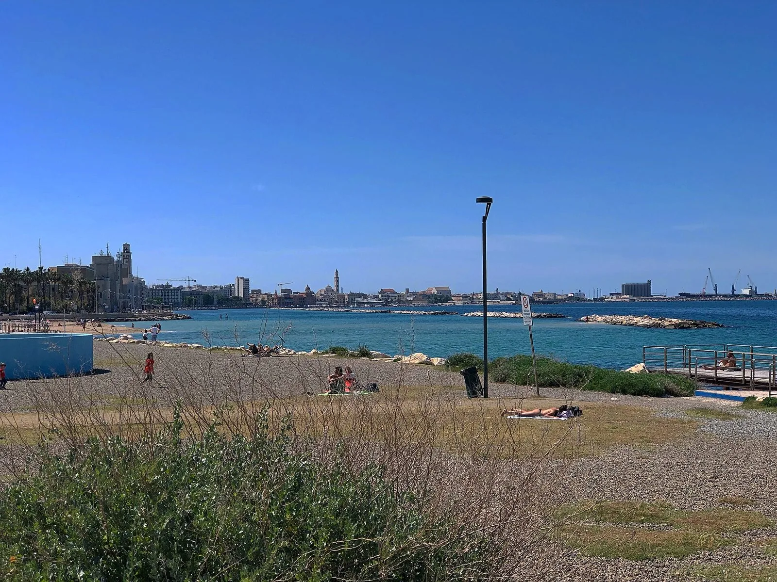 La spiaggia di Pane e Pomodoro a Bari in una giornata di sole, con bagnanti sulla ghiaia, il mare azzurro protetto dai frangiflutti e sullo sfondo il profilo della città