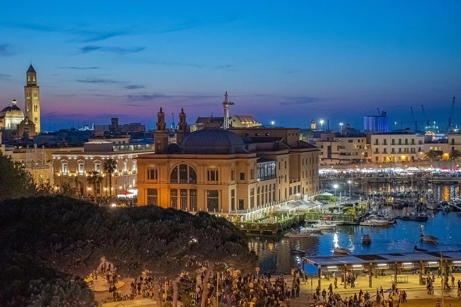 Bari al crepuscolo durante la festa di San Nicola: il Teatro Margherita sul mare illuminato, il porticciolo con le barche, il campanile della cattedrale sullo sfondo e la folla sul lungomare