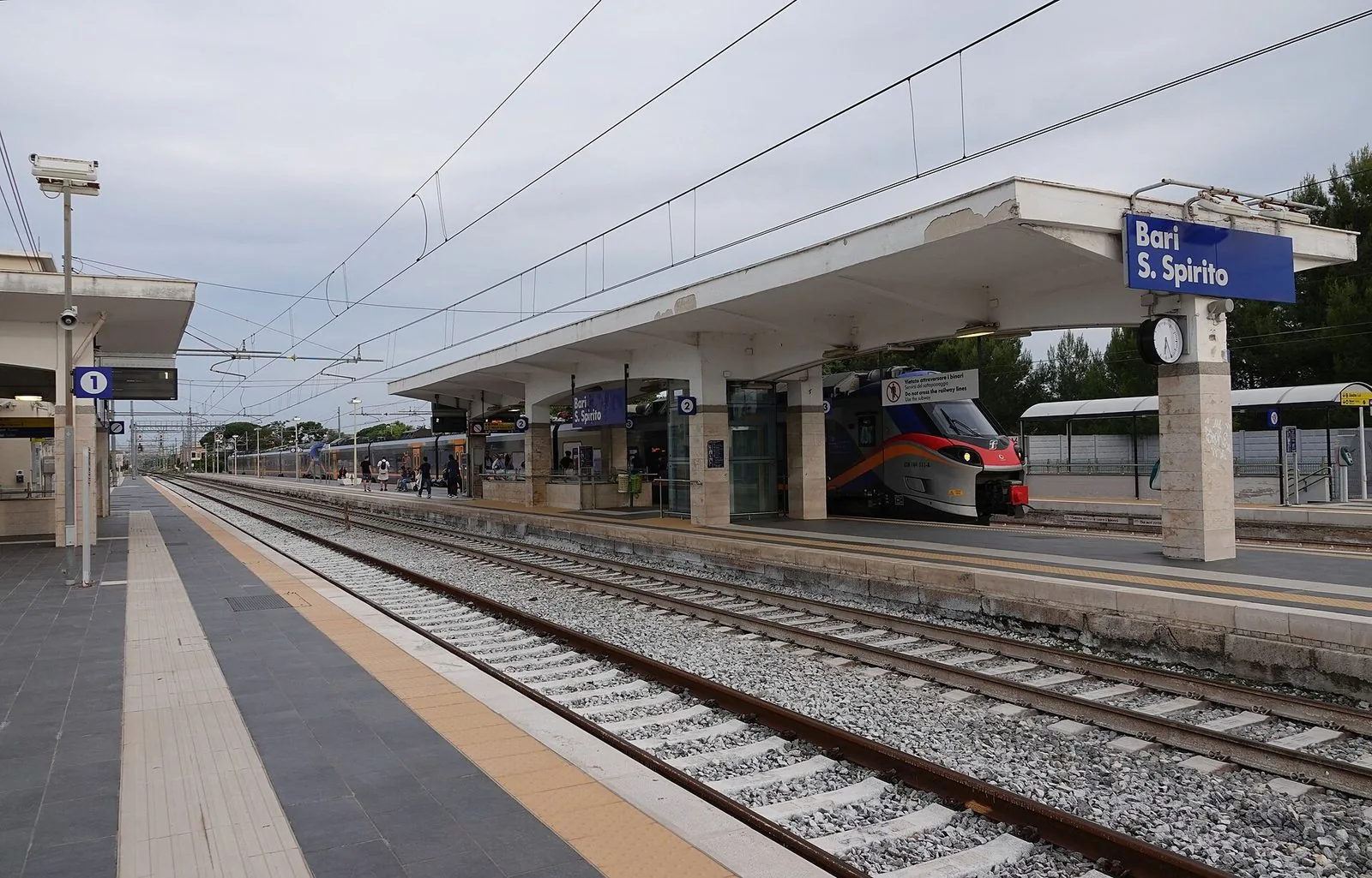 Banchine della stazione di Bari Santo Spirito con il cartello blu, i binari e due treni regionali fermi sotto la pensilina