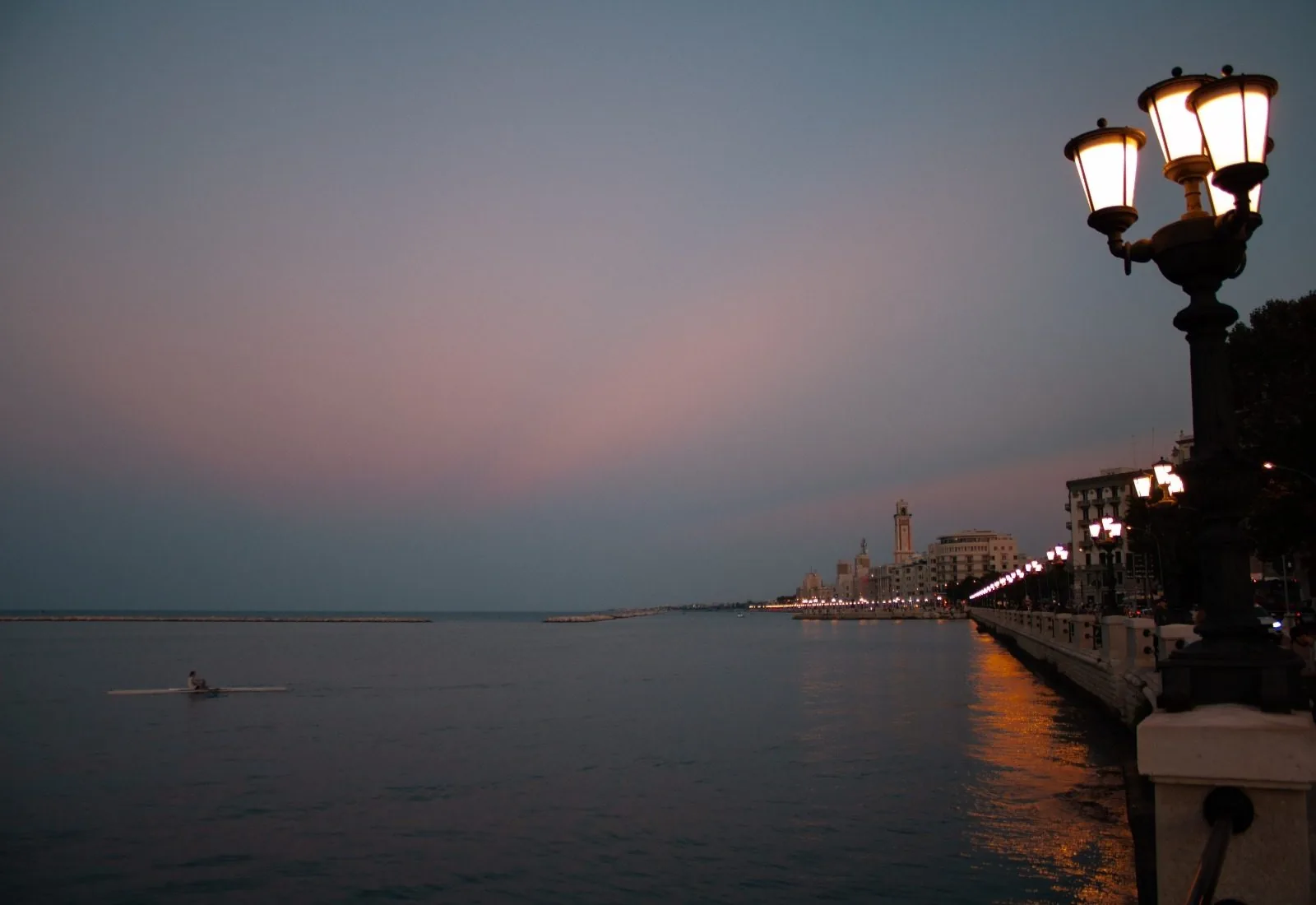 Il lungomare di Bari all'imbrunire, con i lampioni accesi lungo la balaustra, il mare calmo con una piccola barca a remi e in lontananza gli edifici illuminati sulla costa