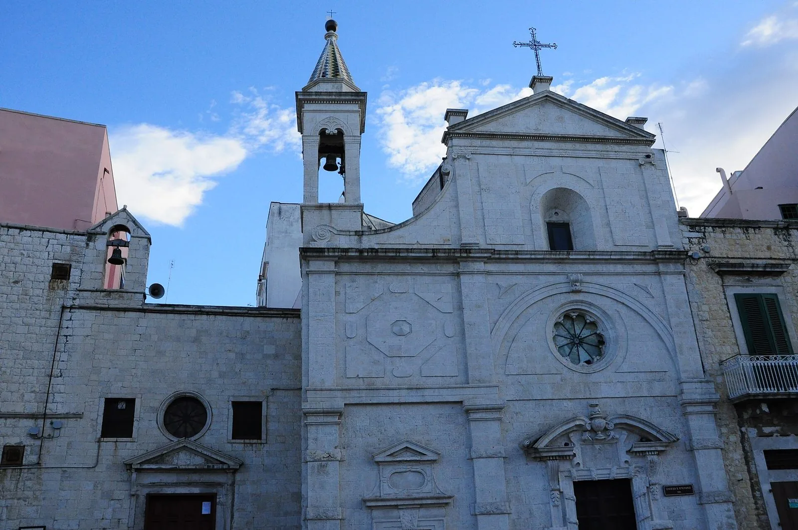 La facciata in pietra bianca della chiesa di Santo Stefano a Molfetta, con il rosone, il portale e il piccolo campanile a cuspide maiolicata contro un cielo azzurro